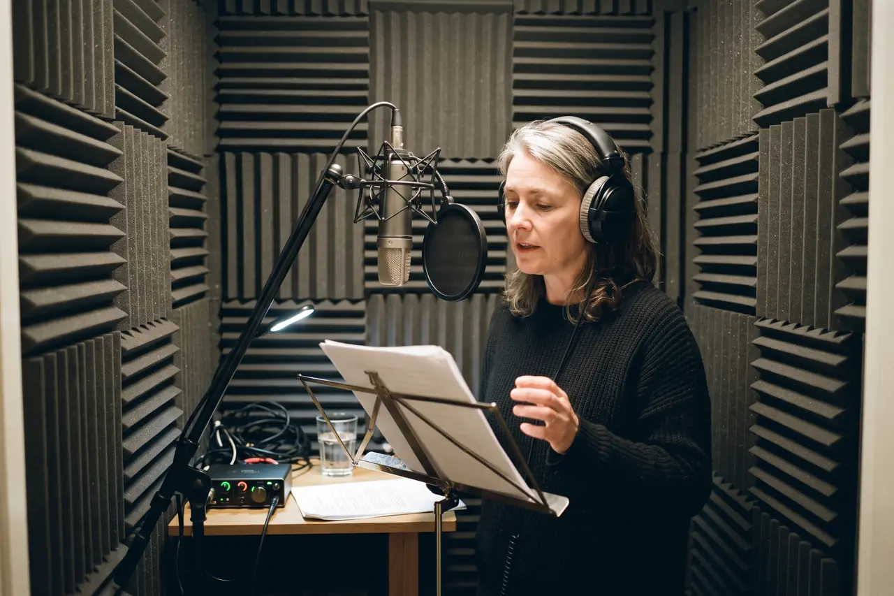 A voice-over artist in headphones reads from a music stand in a small booth lined with acoustic foam, with a condenser microphone and pop filter in front of her